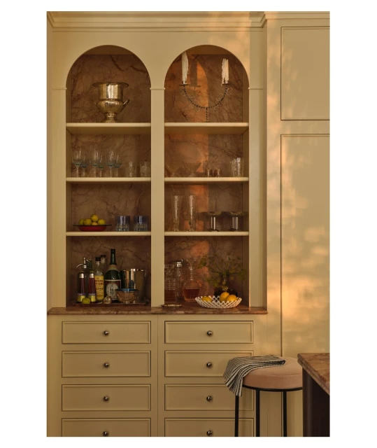 Elegant home bar featuring a built-in cabinet with glasses, bottles, and decorative items, accented by a small upholstered stool.