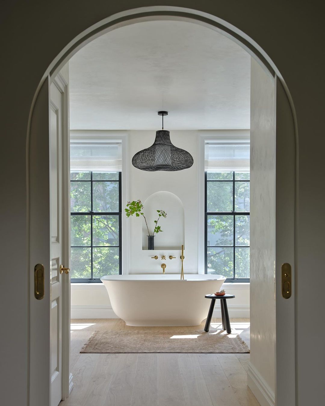 Interior view of a bright bathroom featuring a freestanding bathtub, stool, and dark woven pendant light, framed by a doorway.