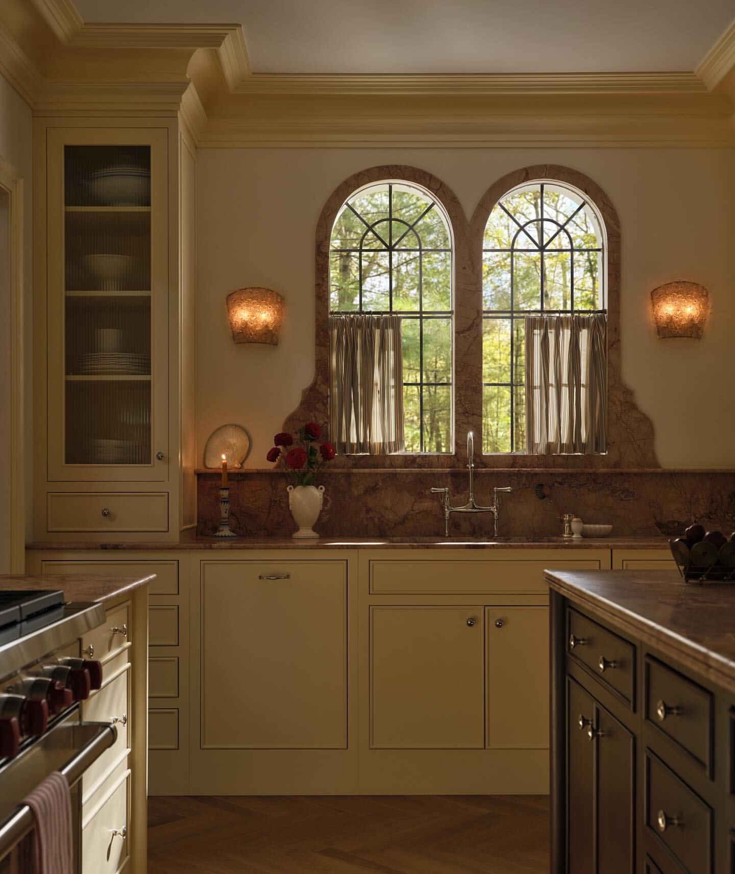 Well-lit kitchen interior with white cabinets, marble countertops, arched windows framing trees, and modern sconces, providing a luxurious ambiance.