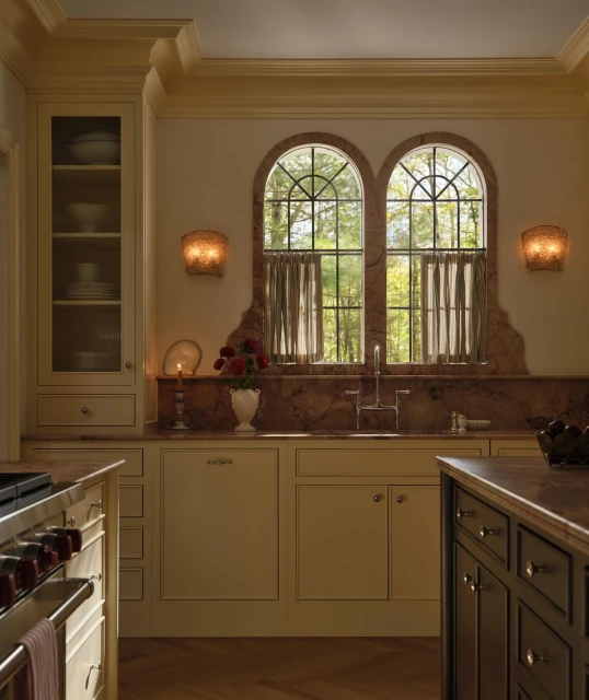 Well-lit kitchen interior with white cabinets, marble countertops, arched windows framing trees, and modern sconces, providing a luxurious ambiance.