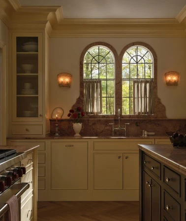 Well-lit kitchen interior with white cabinets, marble countertops, arched windows framing trees, and modern sconces, providing a luxurious ambiance.