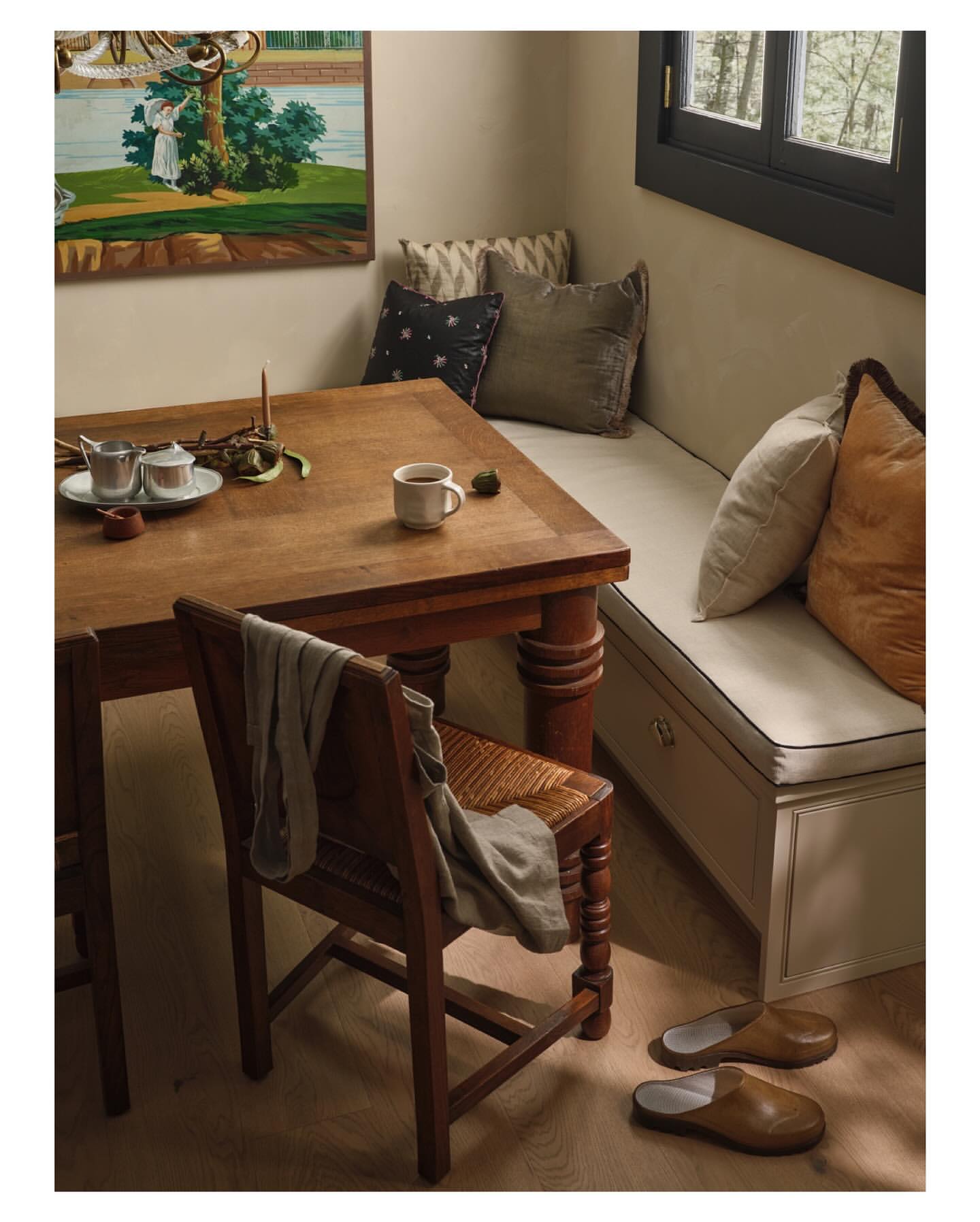 Interior of a cozy dining area with a wooden table, chair, built-in bench, pillows, a painting, and natural light from a window.