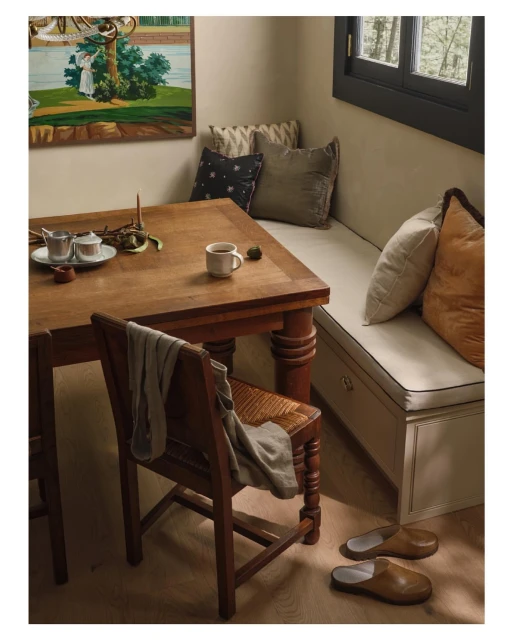 Interior of a cozy dining area with a wooden table, chair, built-in bench, pillows, a painting, and natural light from a window.