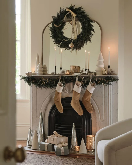 Festive mantelpiece display featuring Christmas stockings, a wreath, candles, and decorative trees, creating a warm, holiday atmosphere.