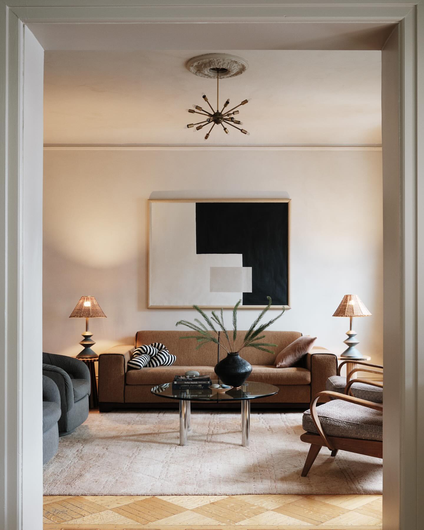 Interior shot of a living room with a sofa, armchairs, round glass table, and graphic wall art in a neutral color palette.