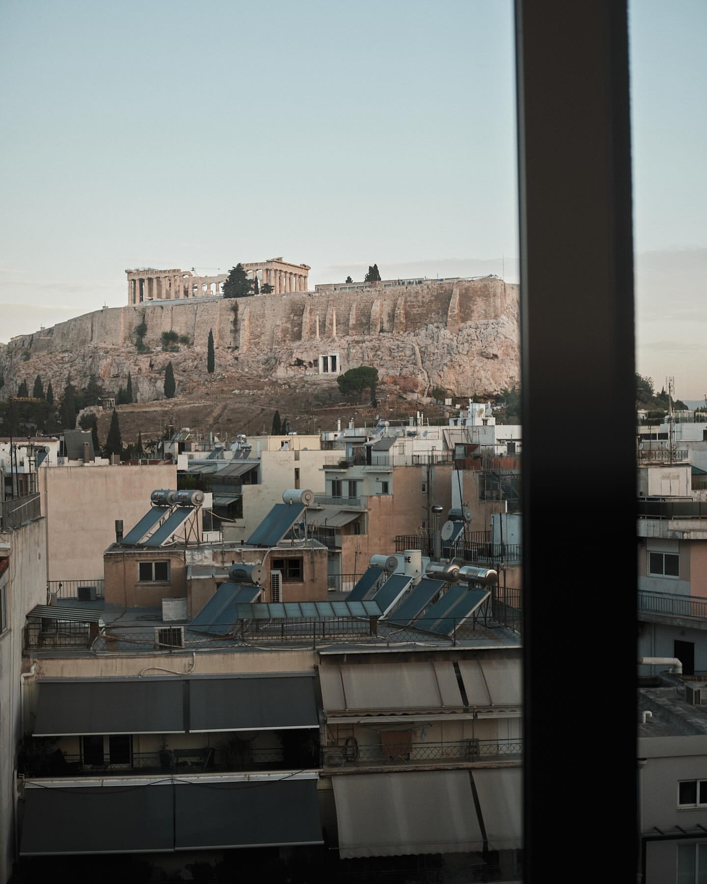 Overlooking Athens, Greece, from a window, featuring city rooftops and the historic Acropolis against a clear sky at daytime.
