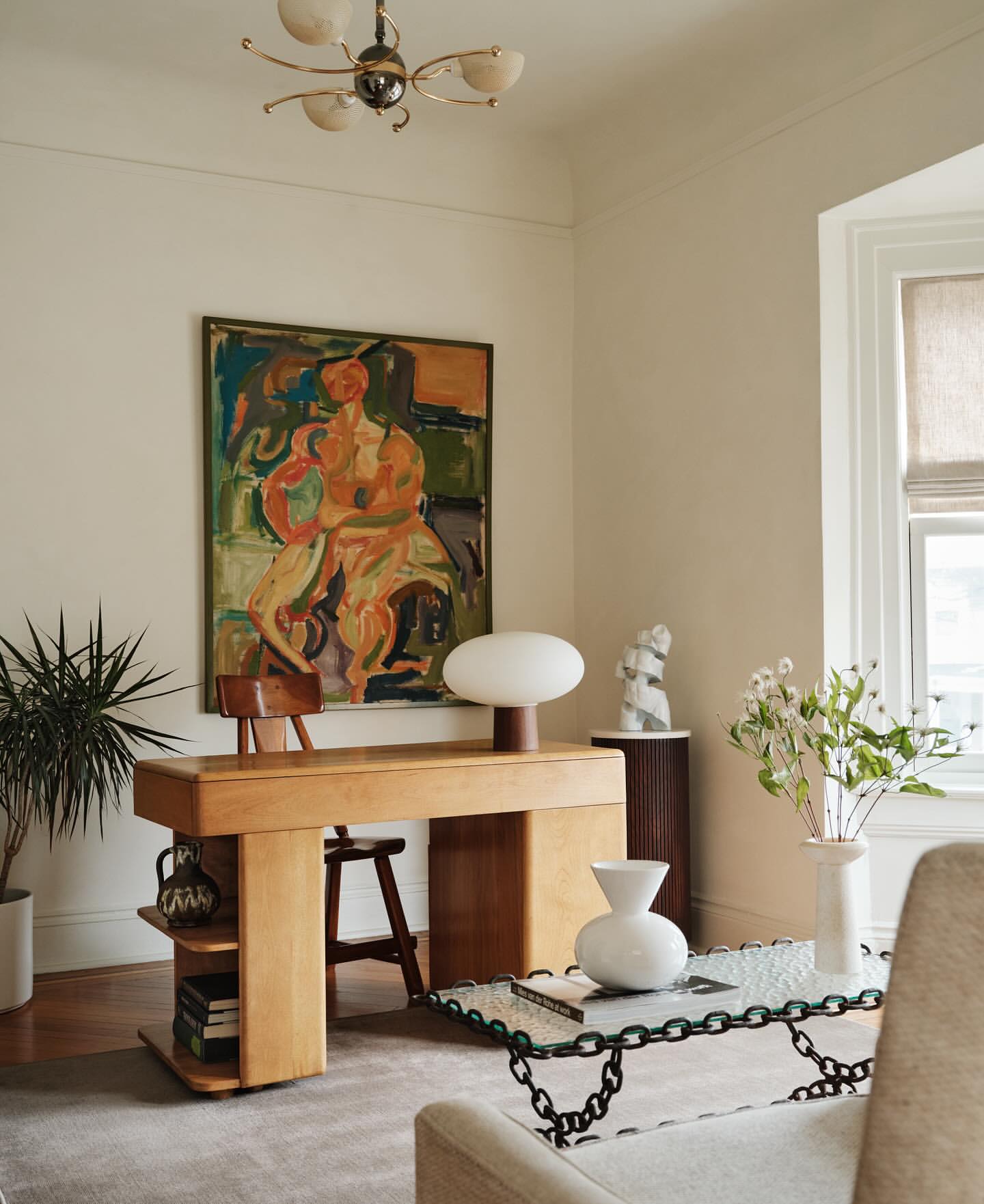 Interior view of living room with blonde desk, modern painting, and white vase with flowers in front of a window.