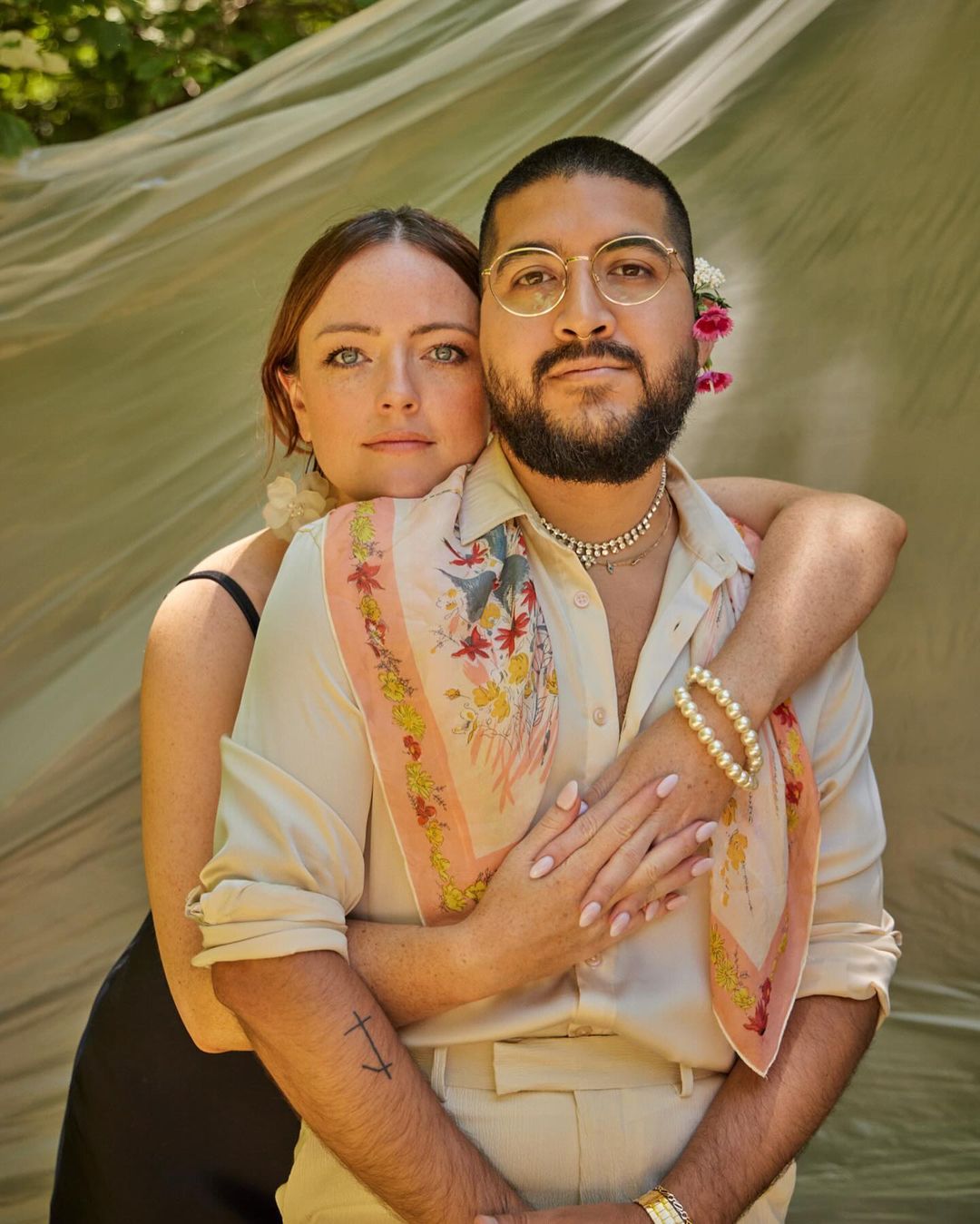 Portrait of a romantic couple embracing outdoors under sheer fabric. The man wears glasses and the woman has her hair styled with flowers.