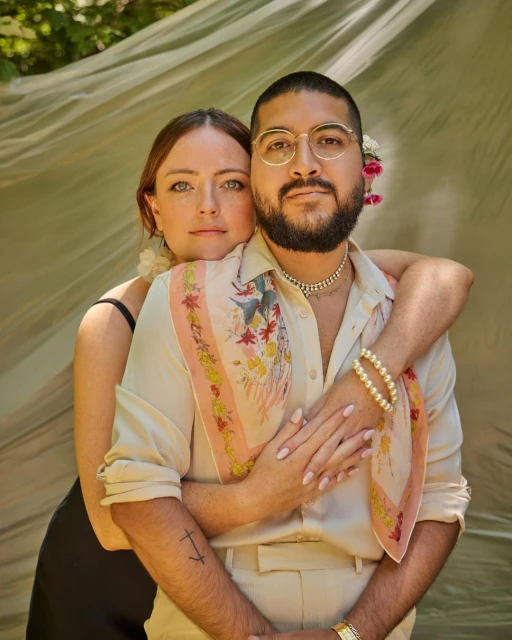 Portrait of a romantic couple embracing outdoors under sheer fabric. The man wears glasses and the woman has her hair styled with flowers.