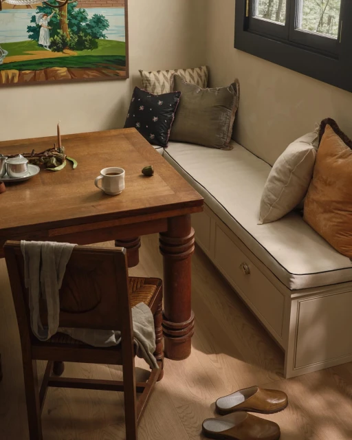 Interior of a home with a wooden table and chair next to a cushioned bench, complemented by decorative pillows and natural lighting.