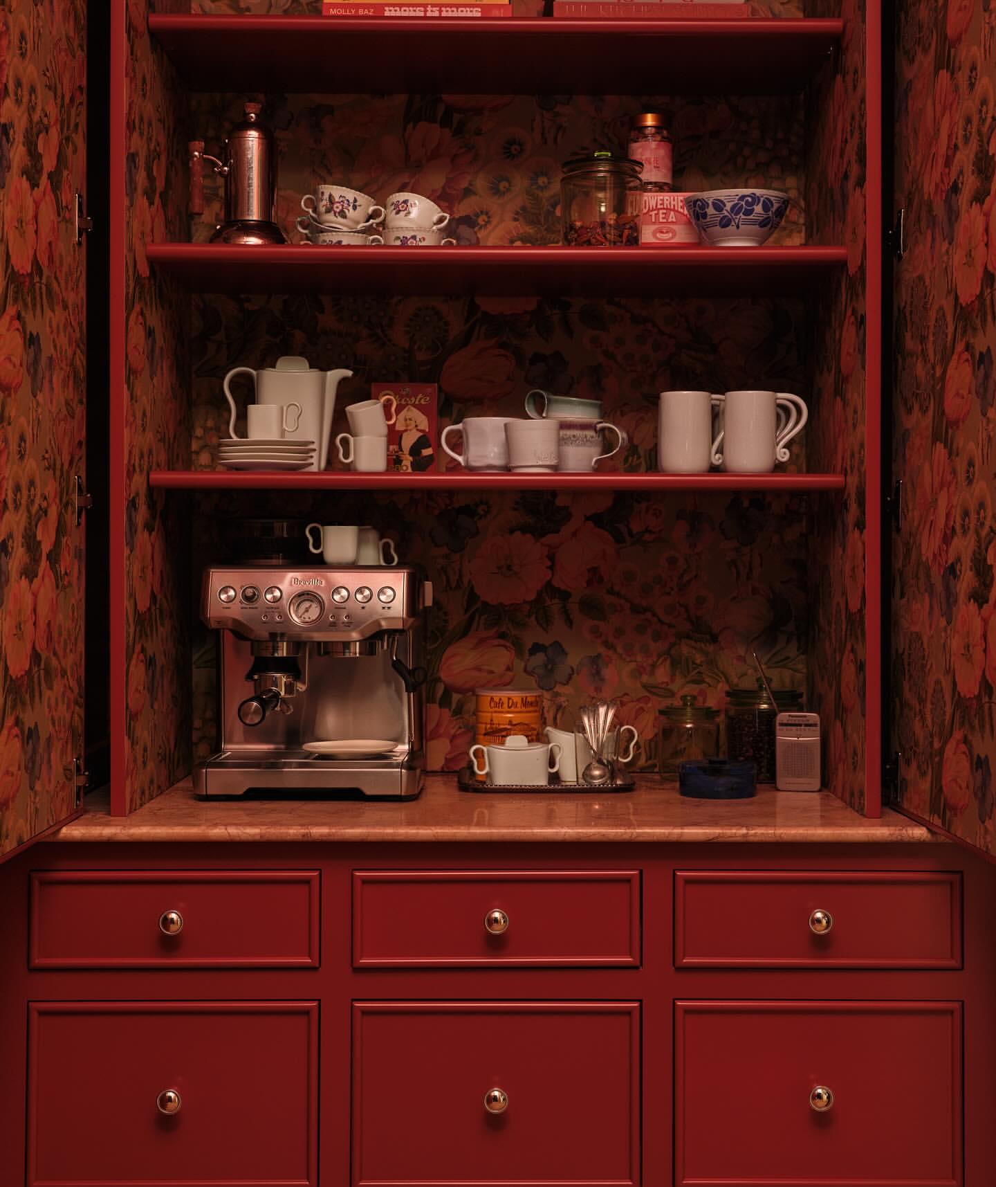 Interior view of a red cabinet with open doors revealing shelves filled with teacups, bowls, an espresso machine, and decorative objects.