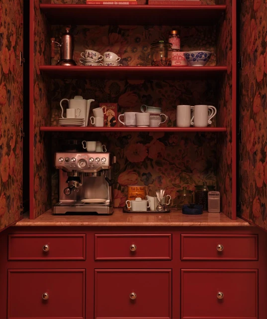 Interior view of a red cabinet with open doors revealing shelves filled with teacups, bowls, an espresso machine, and decorative objects.