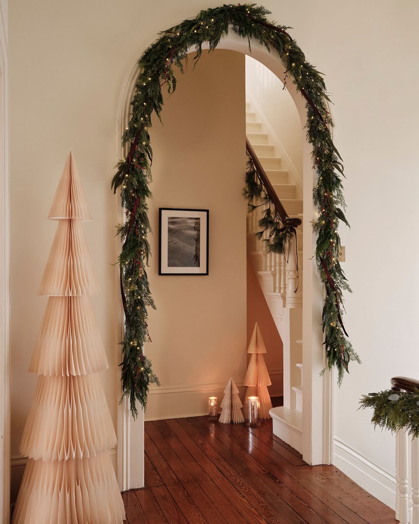 Archway decorated with garland and illuminated with string lights, flanked by paper Christmas trees in a hallway with staircase.