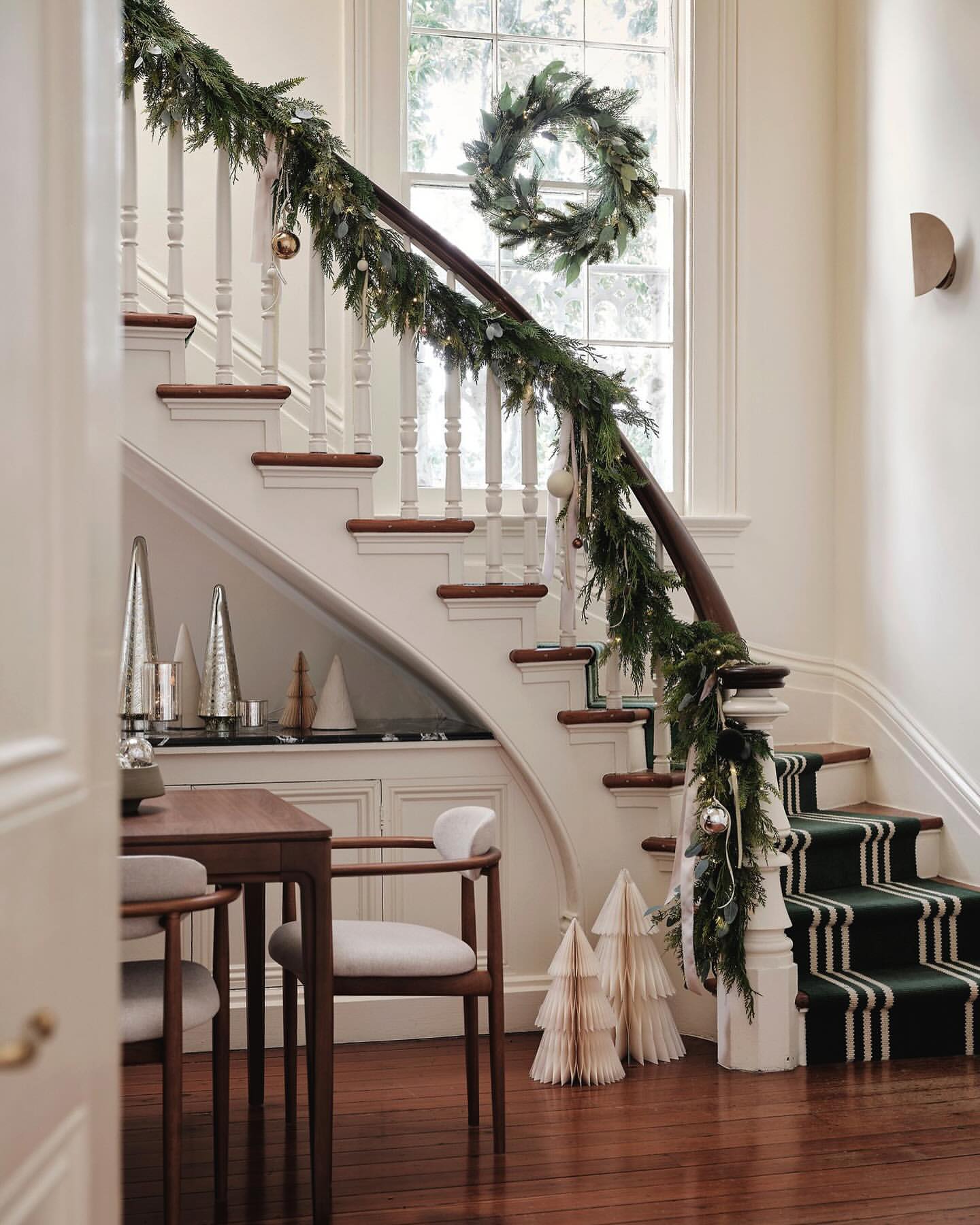 Festive staircase with garland and wreath, Christmas trees on floor, and chairs with table inside home during the holiday season.