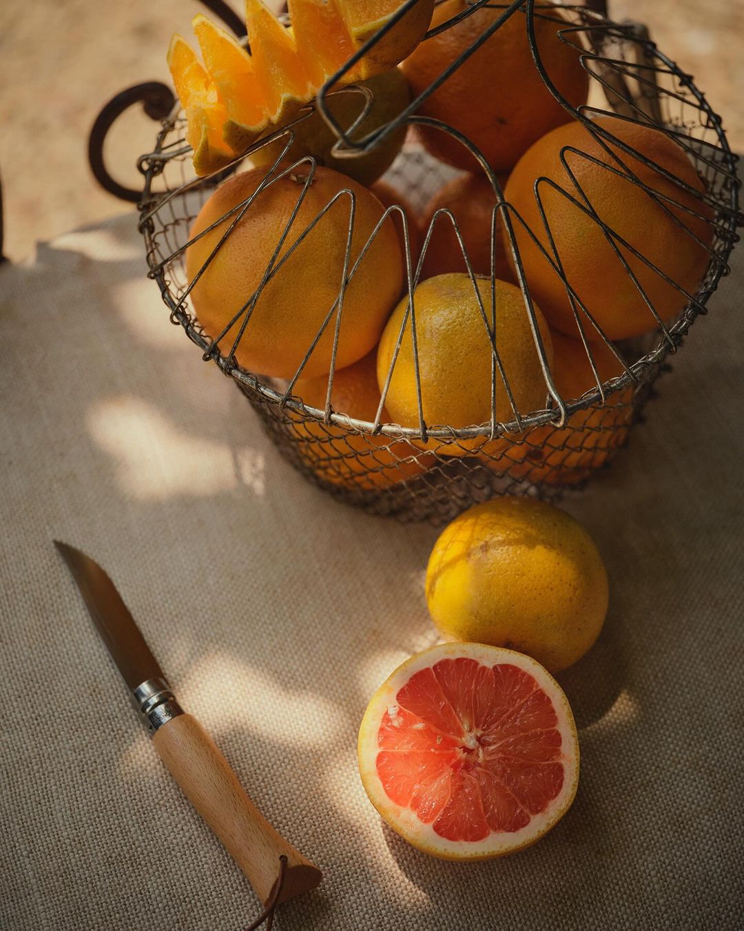 Still life photograph of a basket of oranges with a halved grapefruit and a knife on a linen surface in natural lighting.