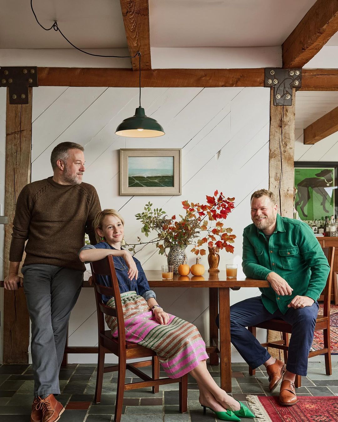 Two men and a woman sitting at a table in a rustic room. Beams cross the ceiling. A painting of a seascape hangs on the wall.