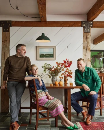 Two men and a woman sitting at a table in a rustic room. Beams cross the ceiling. A painting of a seascape hangs on the wall.