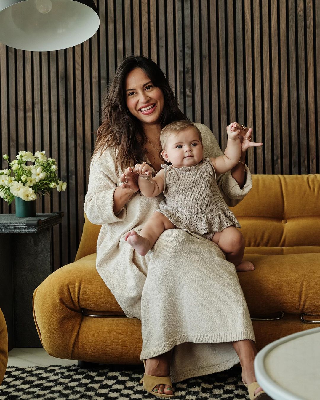 A smiling woman in a beige dress sits with a baby in her lap on a yellow sofa, with a modern wood wall and flowers in the background.