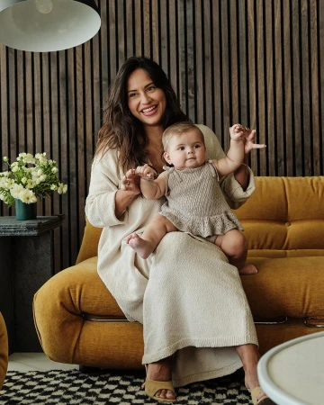 A smiling woman in a beige dress sits with a baby in her lap on a yellow sofa, with a modern wood wall and flowers in the background.