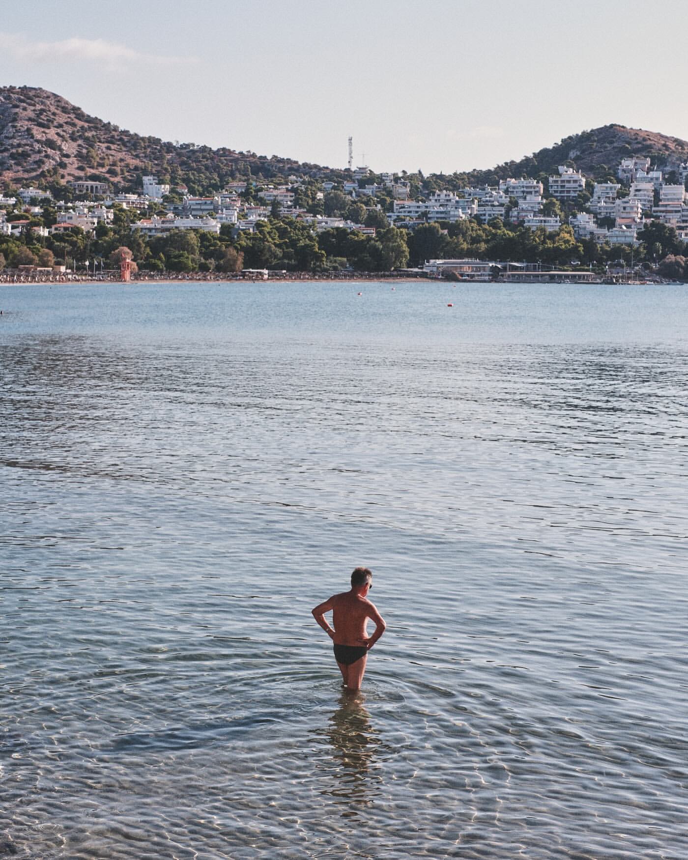 Man in swim trunks standing in the ocean water with mountains and houses visible on the coastline in the background.