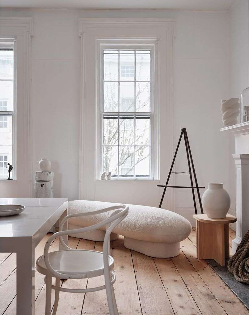 Minimalist room with a white sofa, table, and chair set against a backdrop of white walls, windows, and light wood flooring.