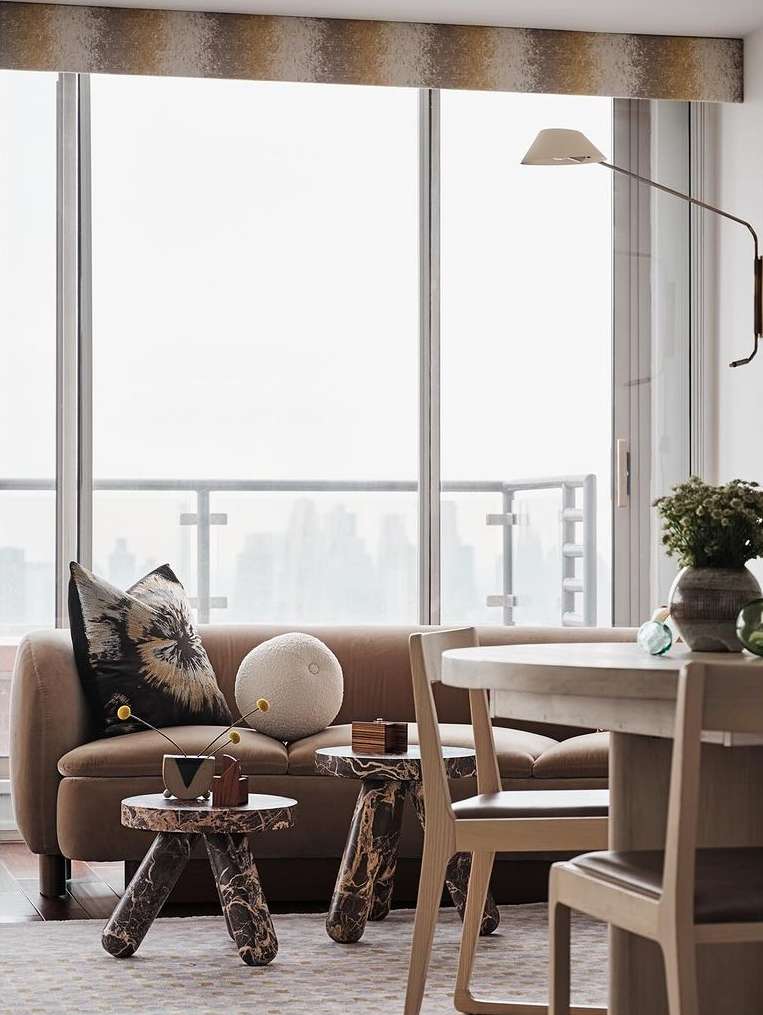 Living room interior with modern furniture: a beige sofa with pillows, two side tables, a dining table set, and a large window.