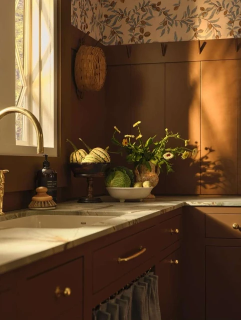 Cozy kitchen interior with brown cabinets, a marble countertop, and a decorative bowl of vegetables and flowers, lit by warm sunlight.