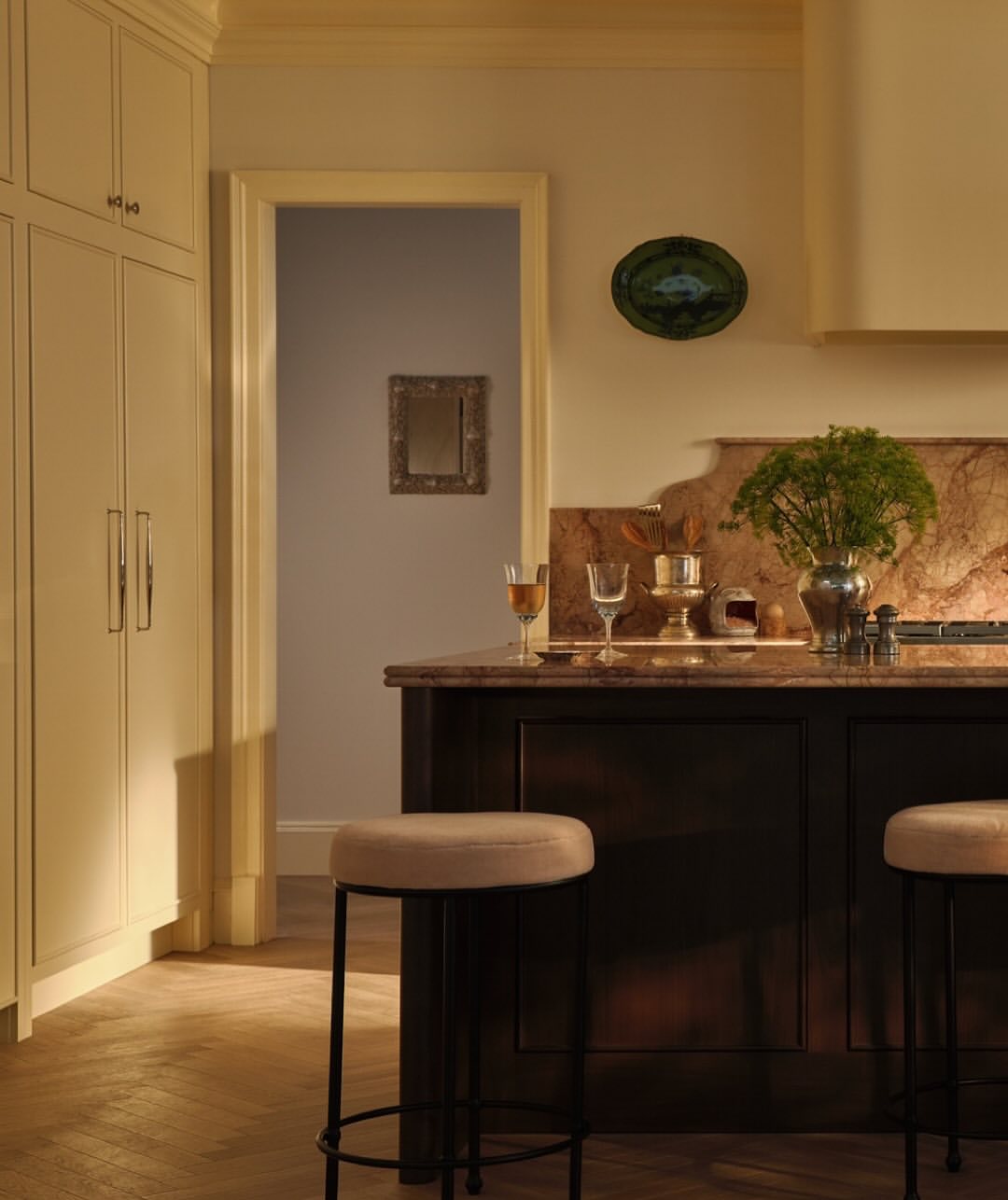 Warm-toned kitchen interior featuring a dark island with stools, marble countertops, and decorative dishware, creating an inviting atmosphere.