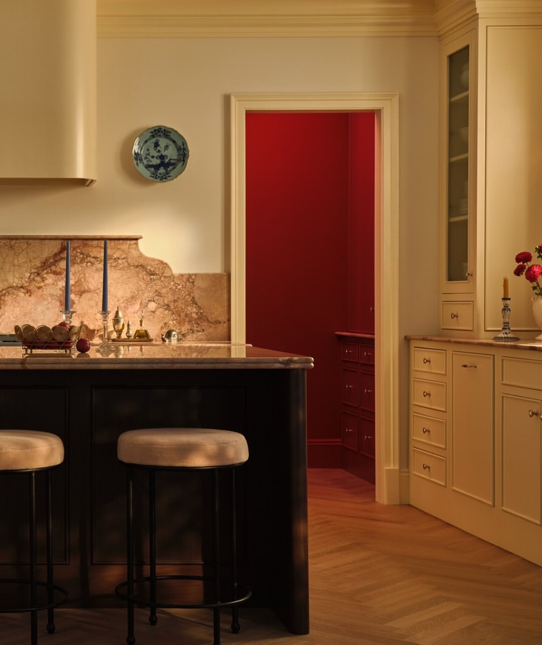 Warm-toned interior view of a kitchen featuring an island with stools, a decorated plate, and an open doorway to a red-walled room.