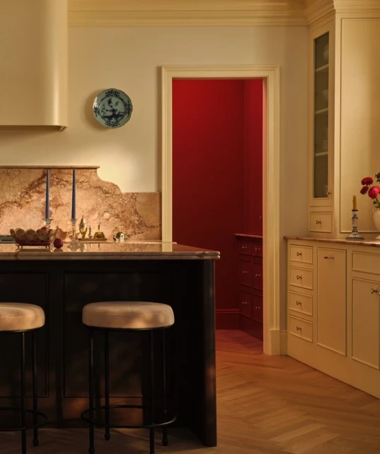 Warm-toned interior view of a kitchen featuring an island with stools, a decorated plate, and an open doorway to a red-walled room.