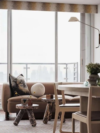Modern living room interior with a beige sofa, patterned cushions, small marble side tables, and a dining set by a large window.