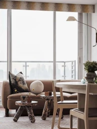 Modern living room interior with a beige sofa, patterned cushions, small marble side tables, and a dining set by a large window.