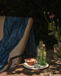 Green glass bottle, small glass, and terracotta bowl on white tray on outdoor wooden table with blue throw draped over lounge chair.