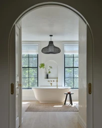 Freestanding white bathtub beneath a dark woven pendant light viewed through an arched doorway