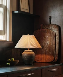 Ceramic table lamp with pleated shade on dark countertop beside antique wooden cutting boards and limes in a bowl
