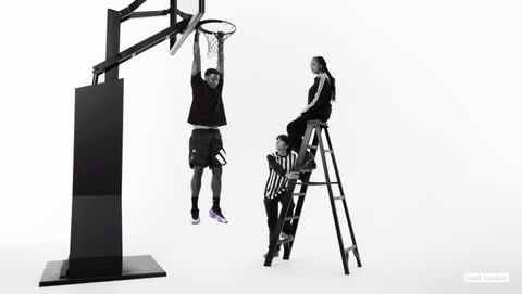 Monochrome shot of basketball player hanging from the hoop as two people stand by on a ladder against a bright white backdrop.