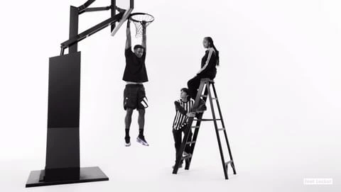 Monochrome shot of basketball player hanging from the hoop as two people stand by on a ladder against a bright white backdrop.