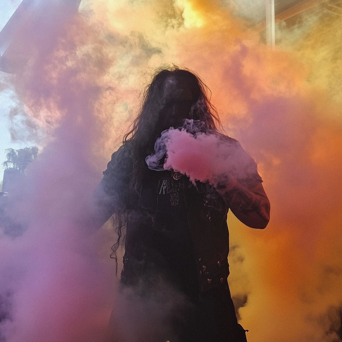 Adult with long dark hair stands outdoors amidst colorful smoke, wearing a black shirt and pants, in a dynamic and artistic composition.