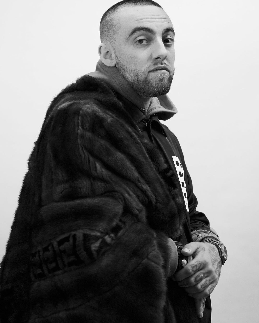 Black and white portrait of an adult male wearing a fur coat and standing against a neutral backdrop, showcasing fashion and style.