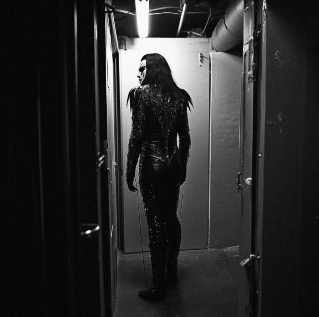 Person in dark costume and makeup stands in dim corridor with white door and overhead light, facing away from camera.