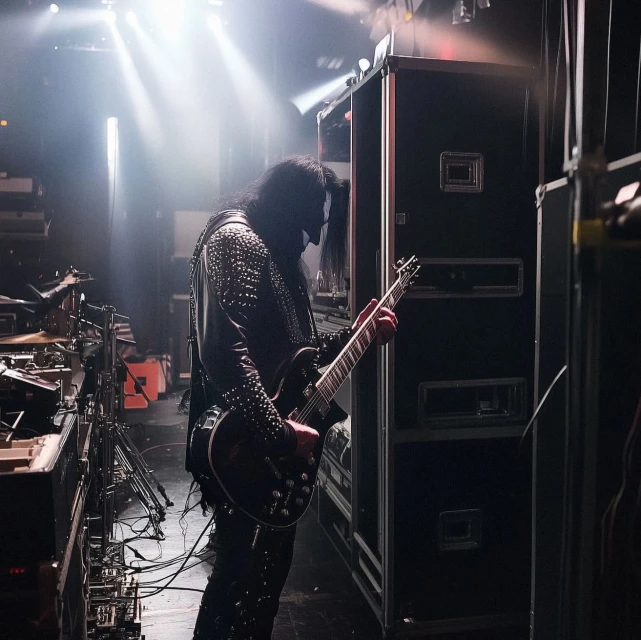 Musician plays black electric guitar on dimly lit stage with cables and speaker cabinets visible in backstage area; concert setting.