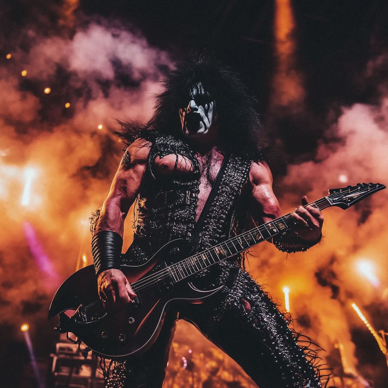 Muscular adult male in stage makeup and elaborate costume plays electric guitar amid bright lights and smoke at a concert venue.