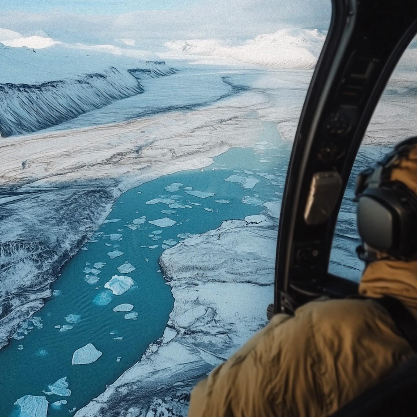 Aerial view from a helicopter over a glacial river filled with icebergs, set against a backdrop of snow-covered mountains in Iceland.