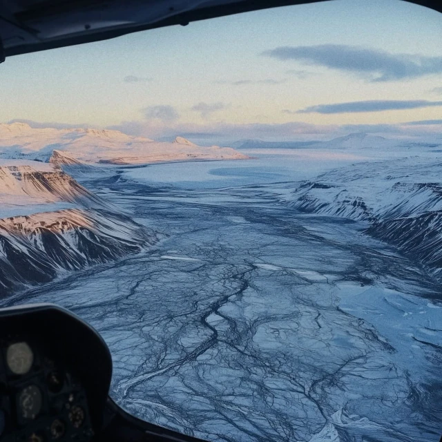Aerial view from inside a helicopter of a snow-covered valley with mountains and a frozen river, showing the cockpit.