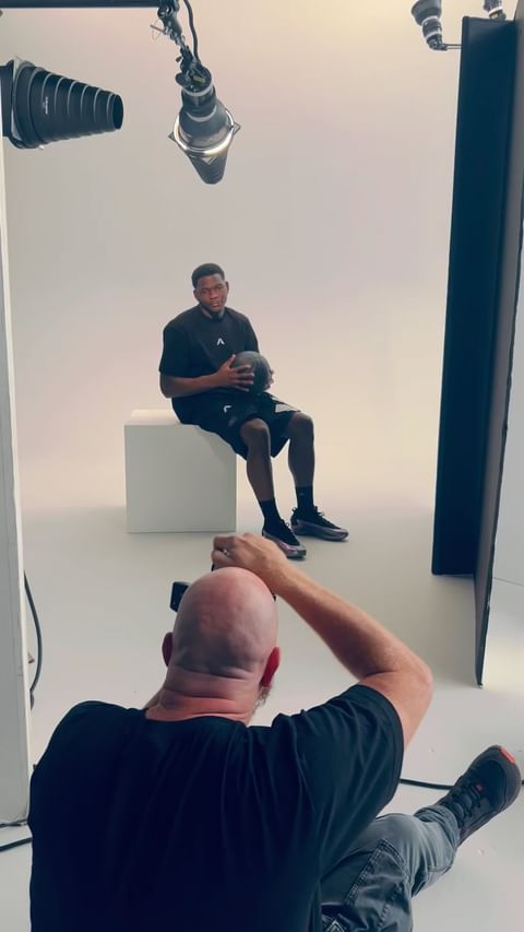 Man in a black t-shirt holds a basketball in a photo studio, seated on a white block. A photographer takes a picture in the foreground.