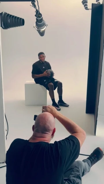Man in a black t-shirt holds a basketball in a photo studio, seated on a white block. A photographer takes a picture in the foreground.