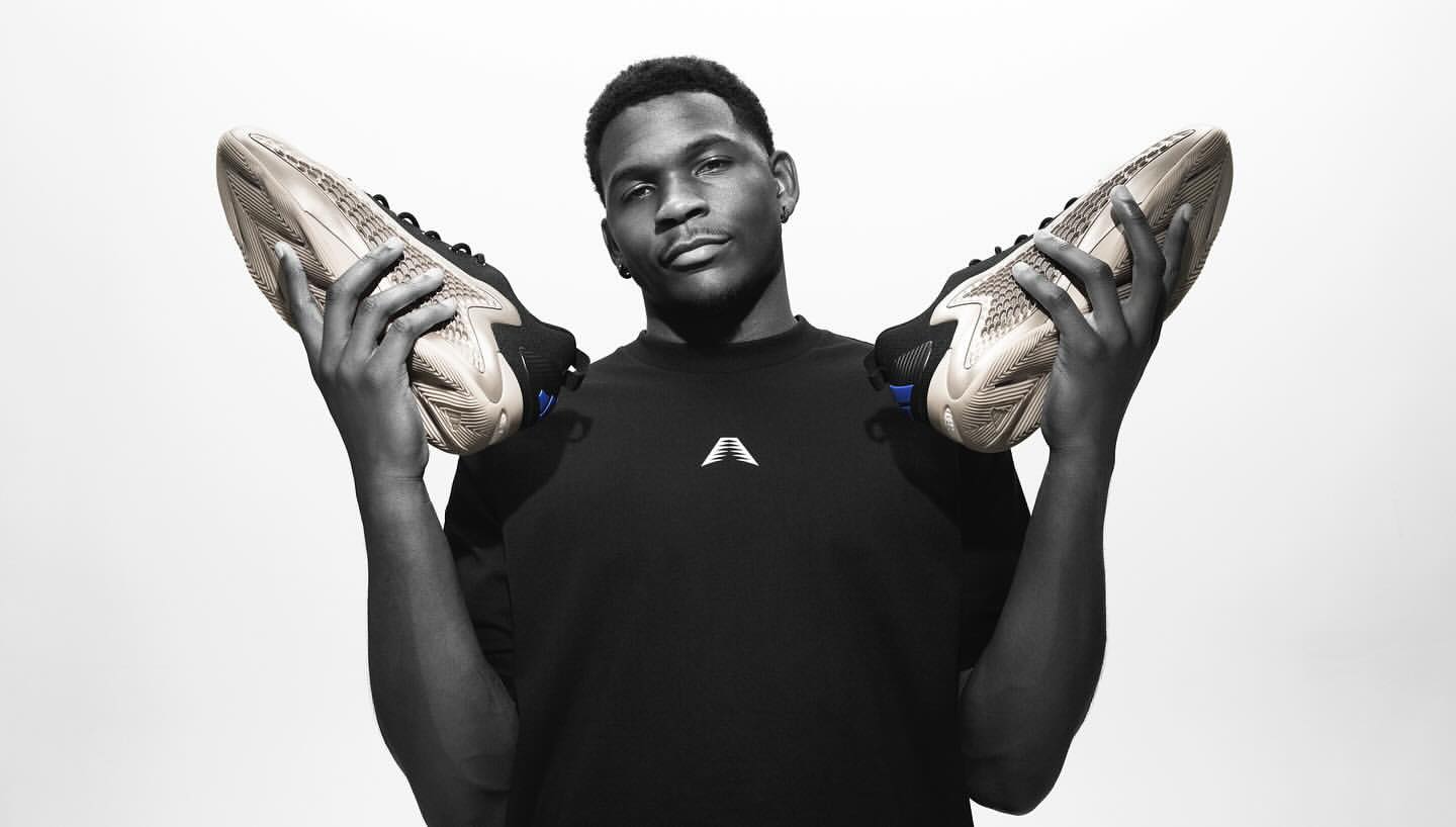 An adult male model holds up two athletic shoes in a black and white studio shot with a bright white background, showcasing footwear.
