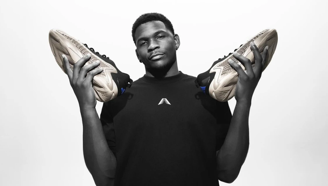 An adult male model holds up two athletic shoes in a black and white studio shot with a bright white background, showcasing footwear.