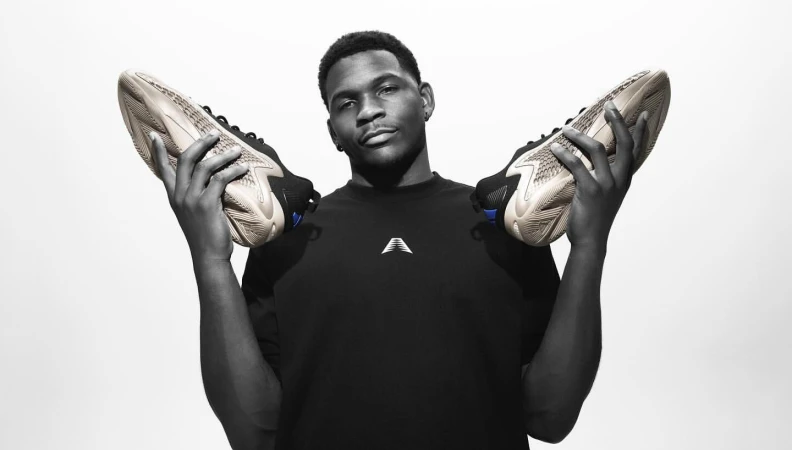 An adult male model holds up two athletic shoes in a black and white studio shot with a bright white background, showcasing footwear.
