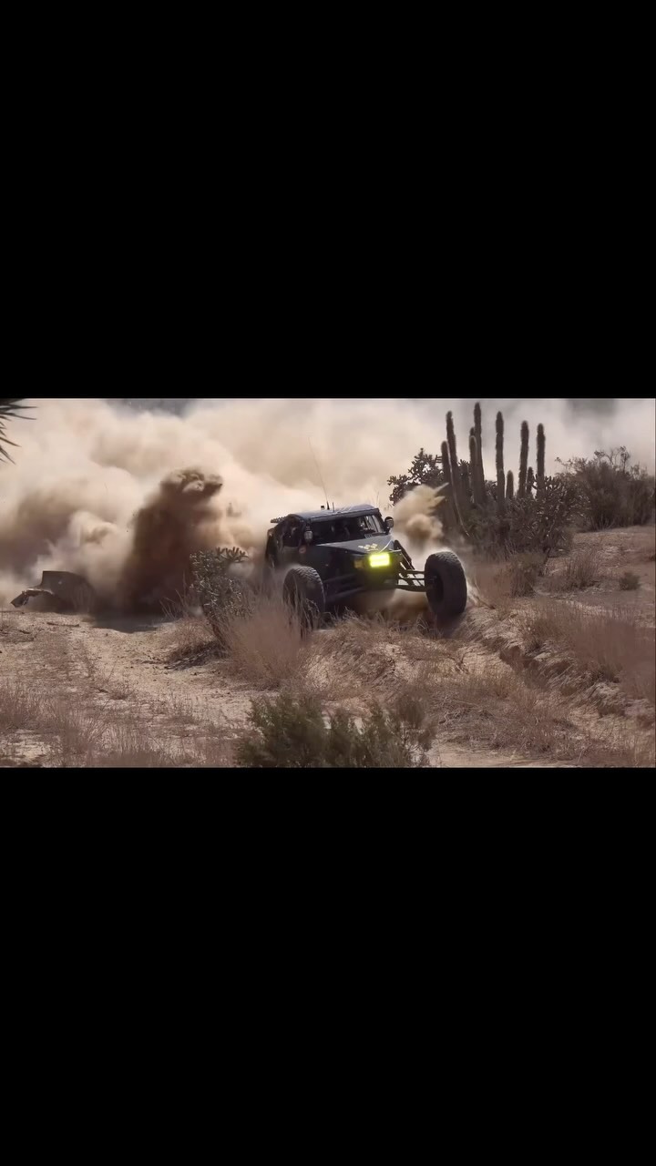 A dark off-road buggy drives through the desert, kicking up a cloud of dust behind it. Cacti are visible in the background.
